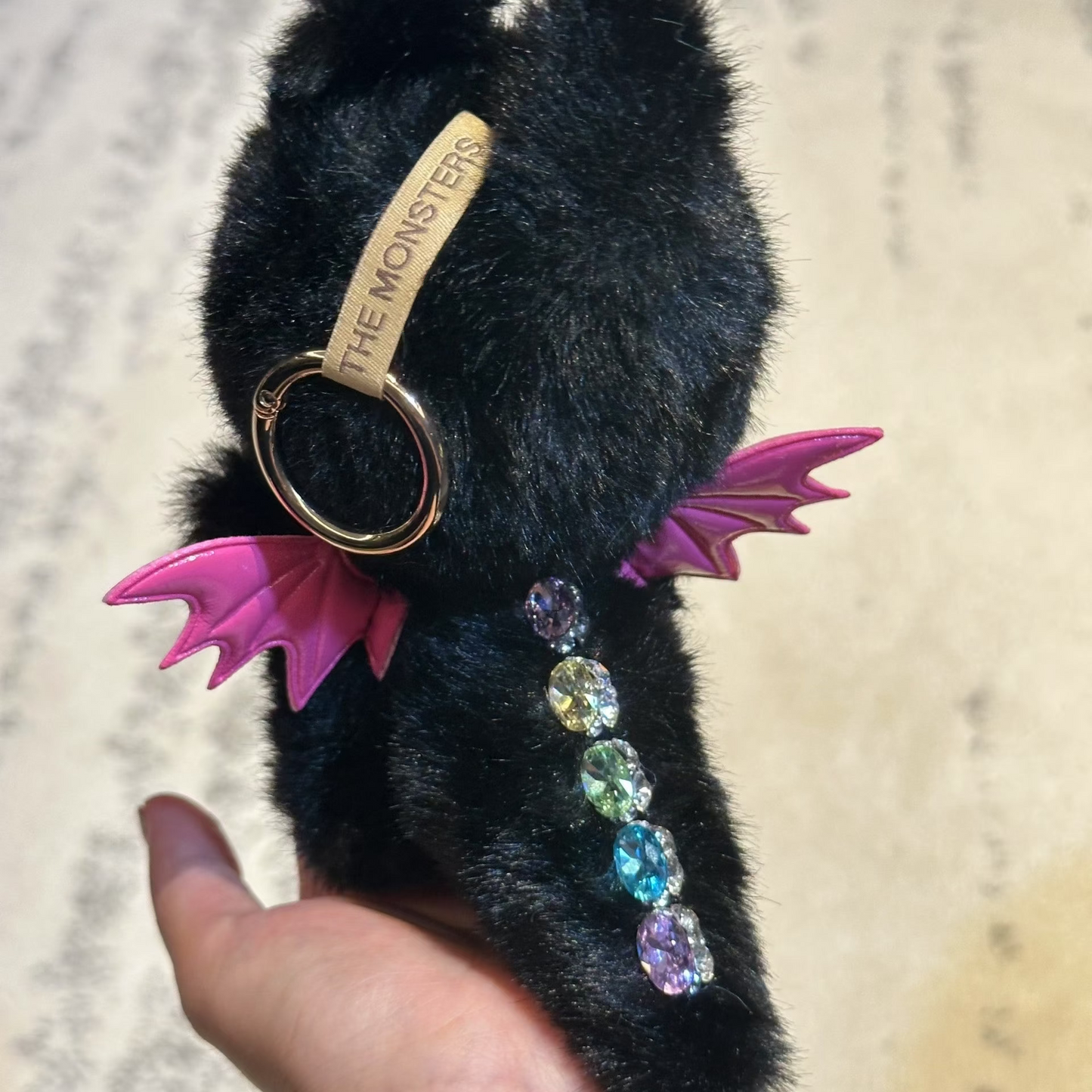 Black plush keychain with pink wings and colorful beads held by a hand against a neutral background