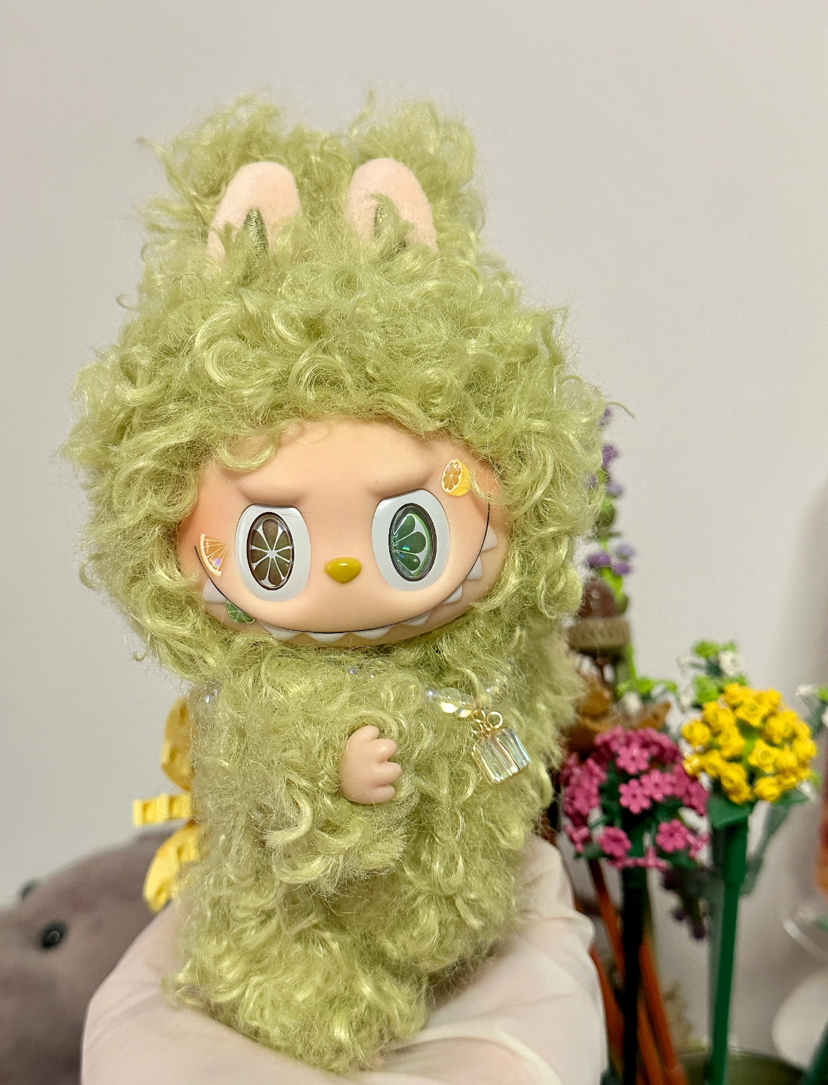 Green plush toy with bunny ears on a white surface with flowers in the background