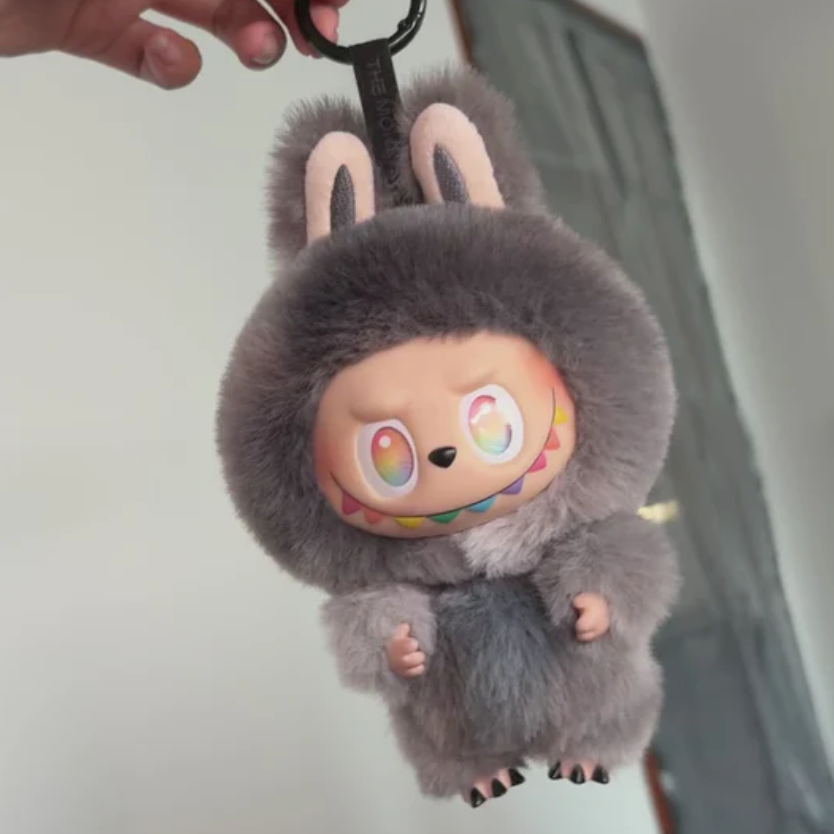 A plush character keychain or bag charm being held by a hand. The character has a fuzzy gray body and hood with pink inner ears, a peach face, large colorful eyes, and a black key ring at the top.