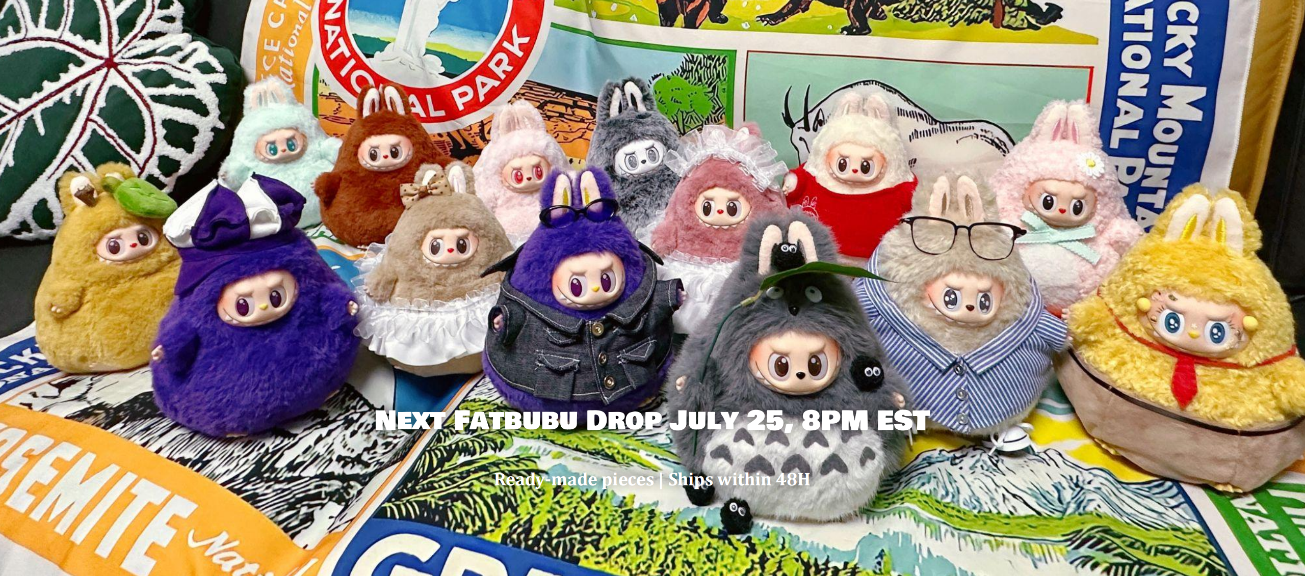 A group of plush character toys arranged together on colorful printed fabric, with a white banner across the center containing text about a drop date. The toys have cartoon faces and wear various outfits in colors like purple, gray, yellow, pink, and red.